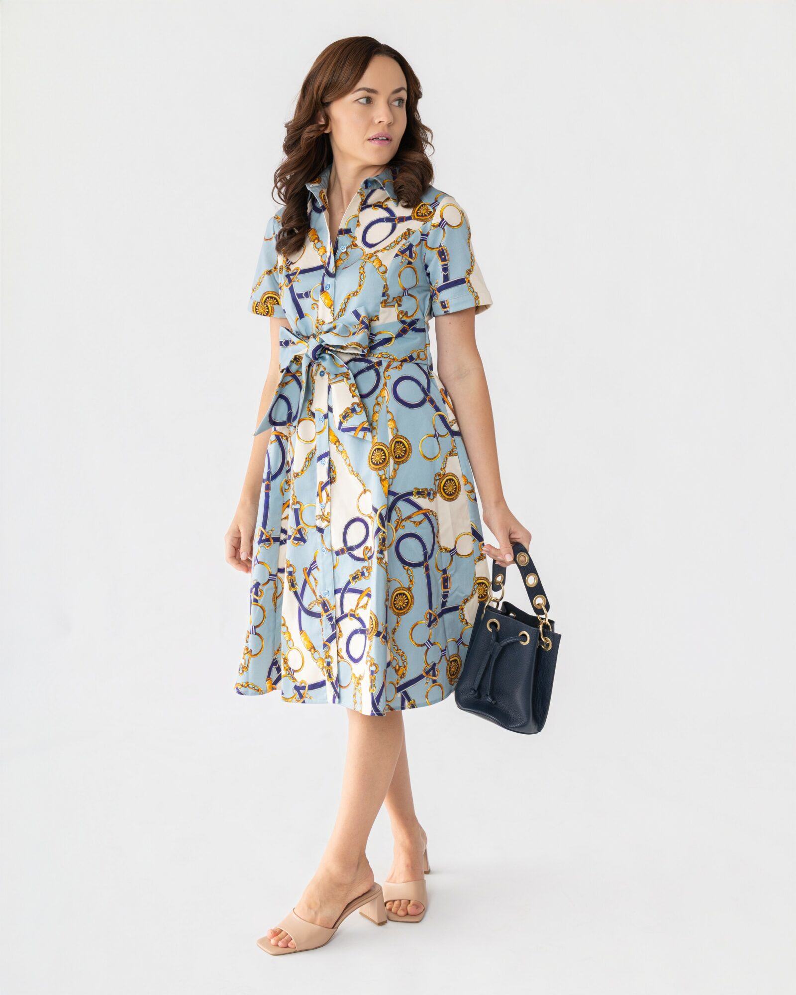 Woman in a patterned dress holding a handbag, standing against a plain background.