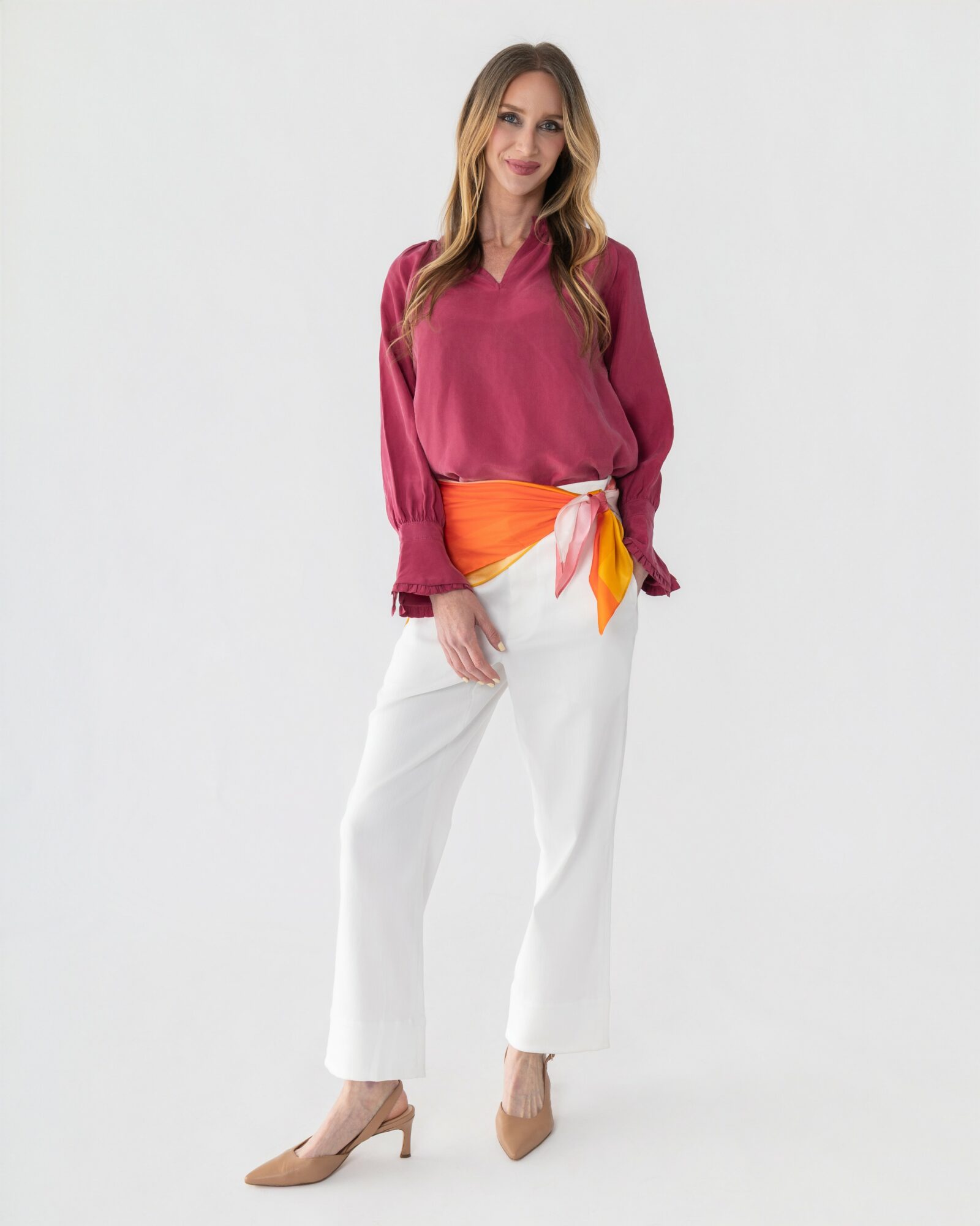 Woman with long hair wearing a pink blouse, white pants, and beige heels, standing against a plain white background.