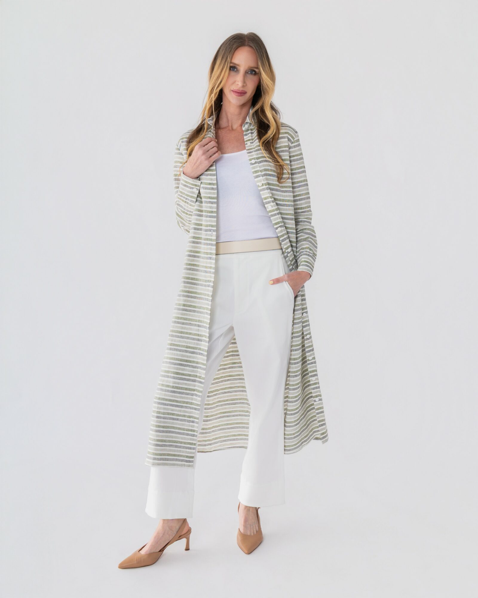 Woman with long hair in a striped long cardigan, white top, white pants, and beige heels, standing against a plain background.