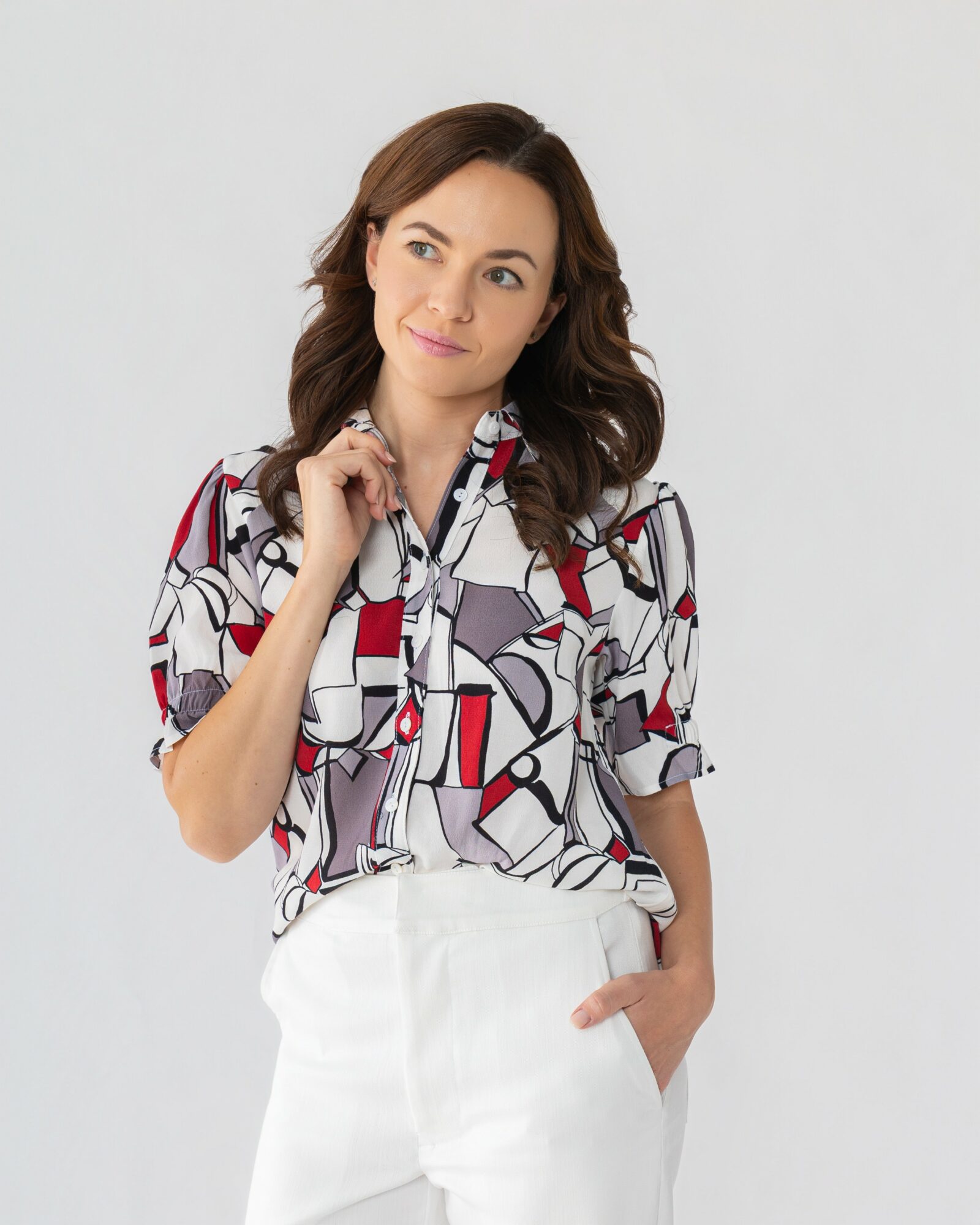 Woman with brown hair wearing a patterned blouse and white pants, standing against a plain background.