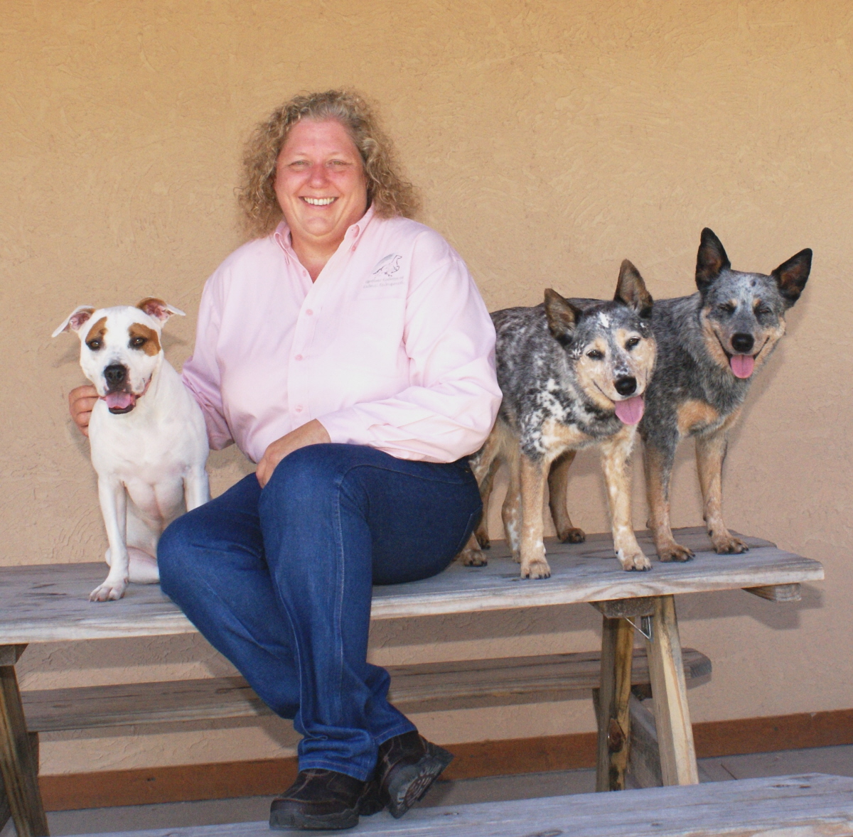 Woman sitting on a wooden bench with three dogs, smiling, against a plain wall.