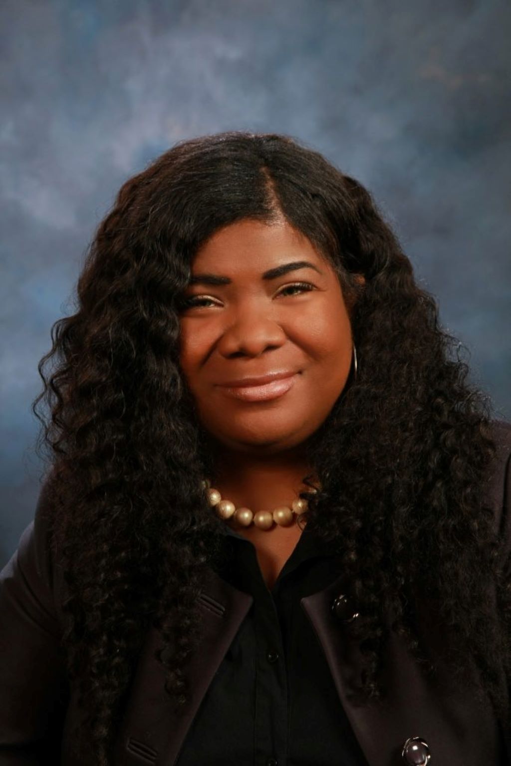 Portrait of a woman with long, curly hair wearing a pearl necklace and dark clothing, smiling softly.