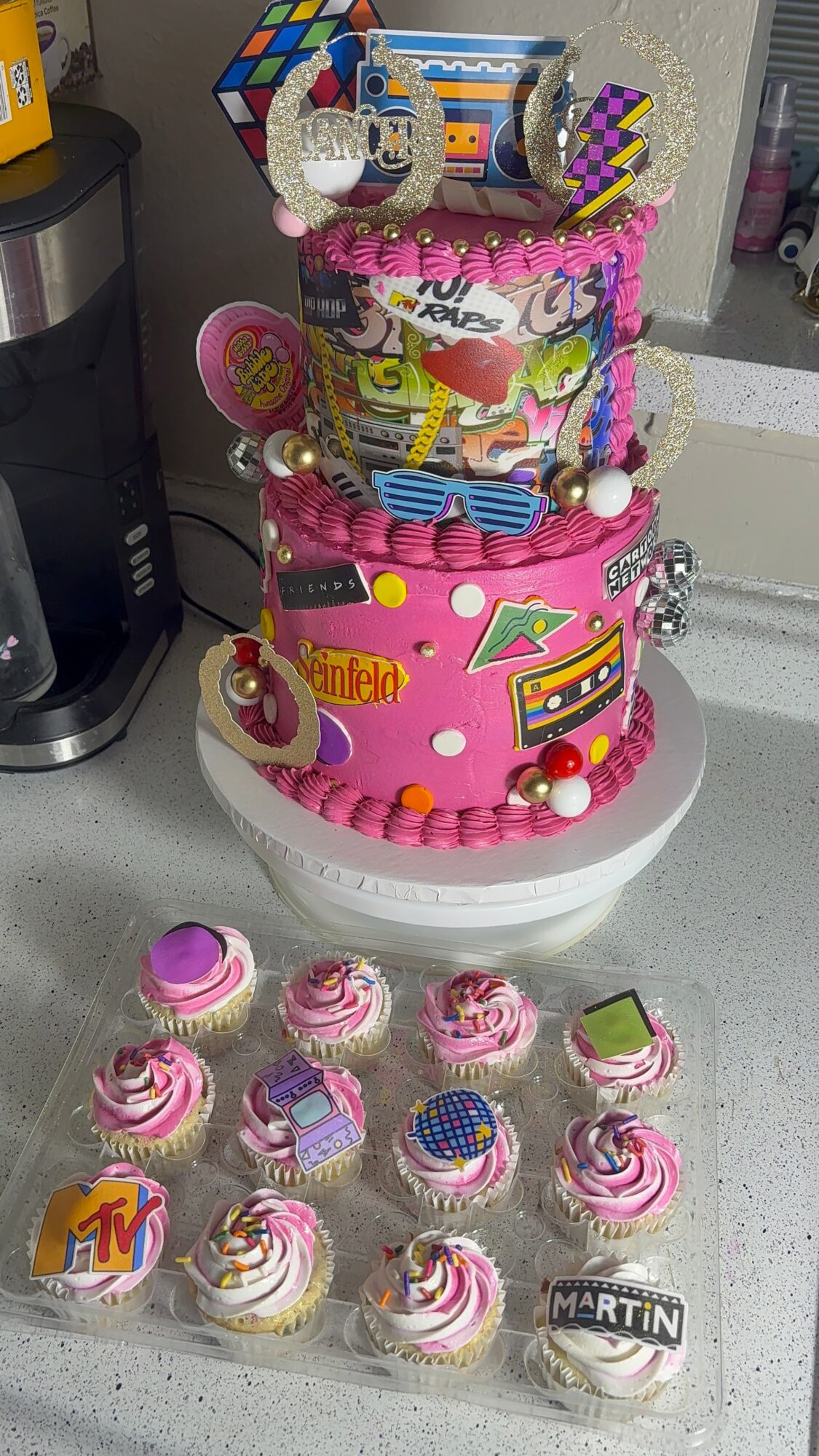 Three-tiered cake decorated with colorful candies, toys, and a pink theme, surrounded by cupcakes with pink and purple frosting.