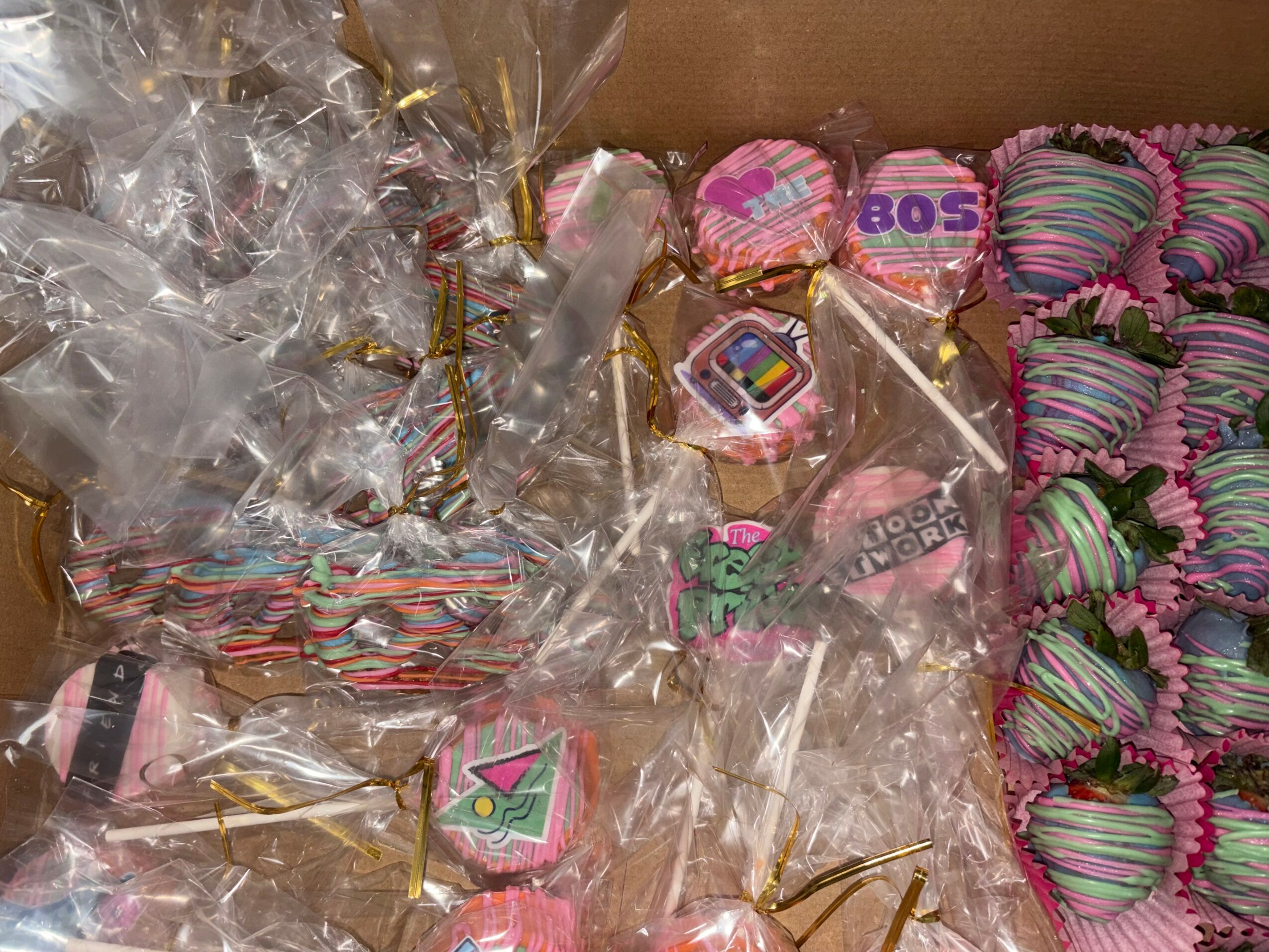 Colorful decorated candies and treats in clear plastic wrappers, arranged in a box, with pink, purple, and green stripes, and various designs.