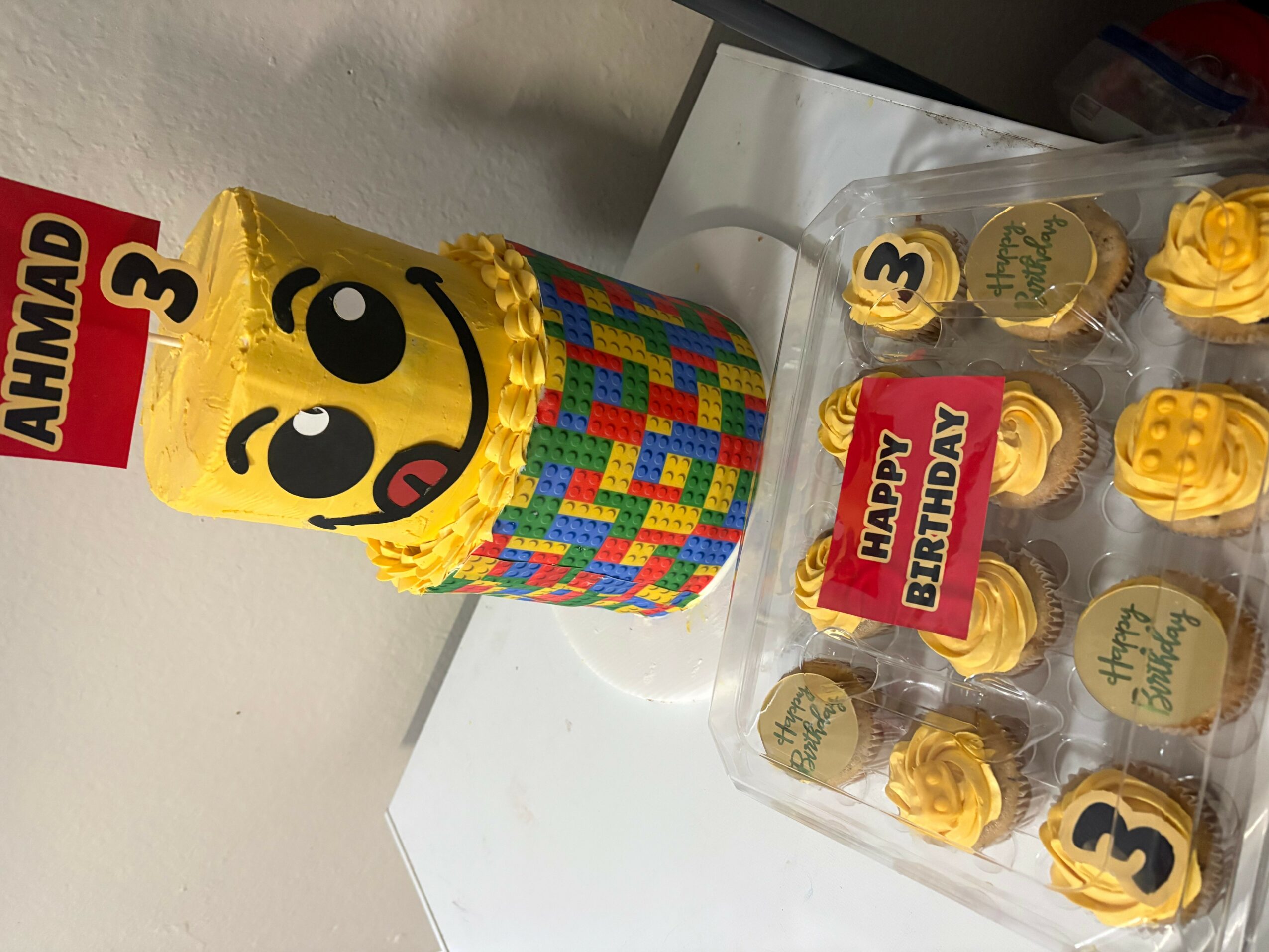 Colorful birthday cake with a smiling face, surrounded by cupcakes with yellow frosting and number toppers, on a white surface.