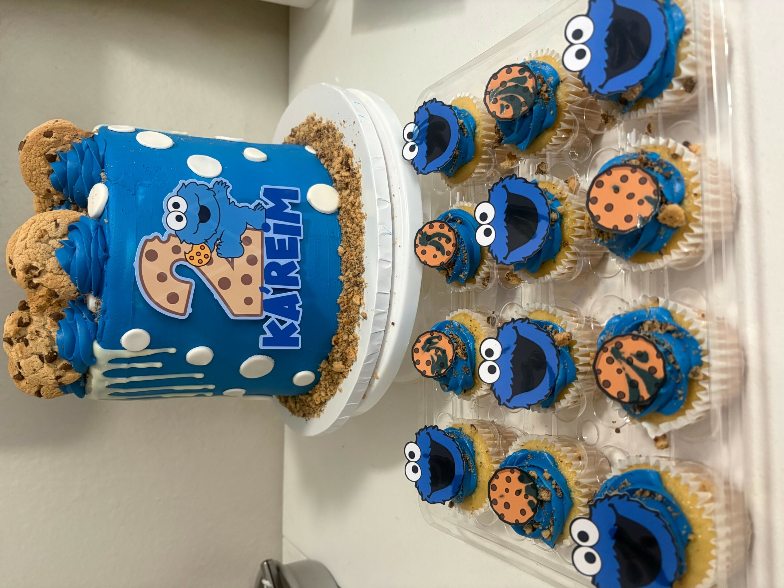 Birthday cake with blue icing and cookie decorations, surrounded by cupcakes with blue and orange icing, Sesame Street theme, number 2 topper.