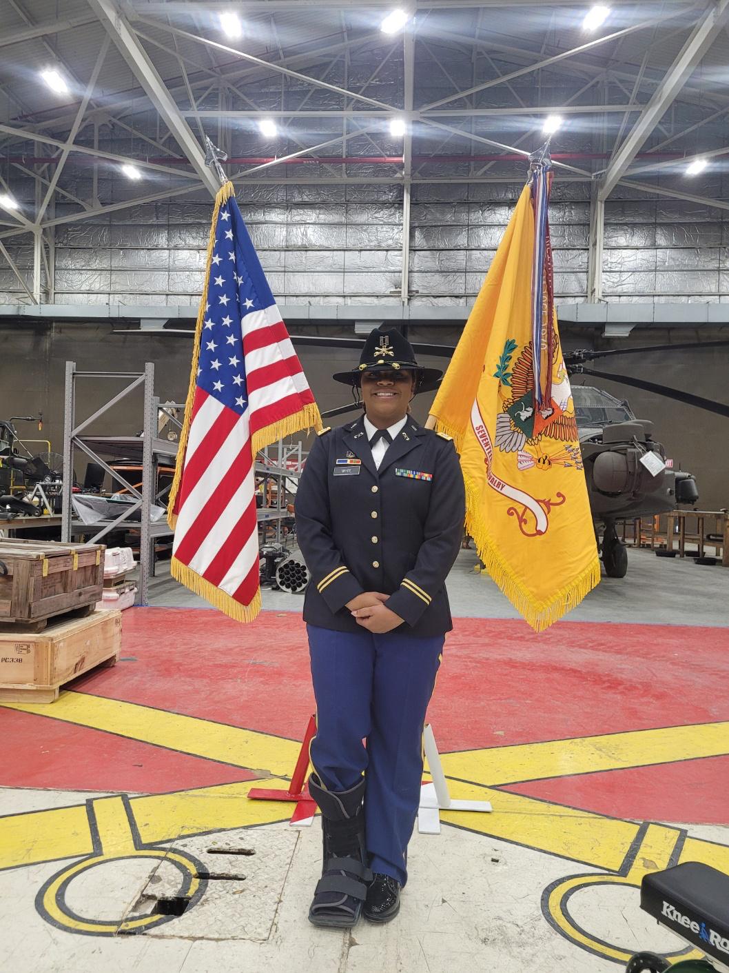 A person in military uniform standing between American and military flags inside a hangar, with aircraft and equipment in background.