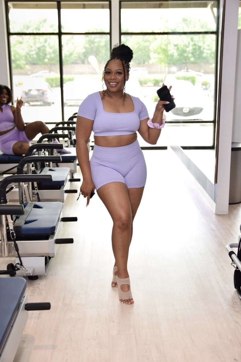 Woman in workout outfit holding a phone, smiling, in a gym with large windows and exercise equipment.
