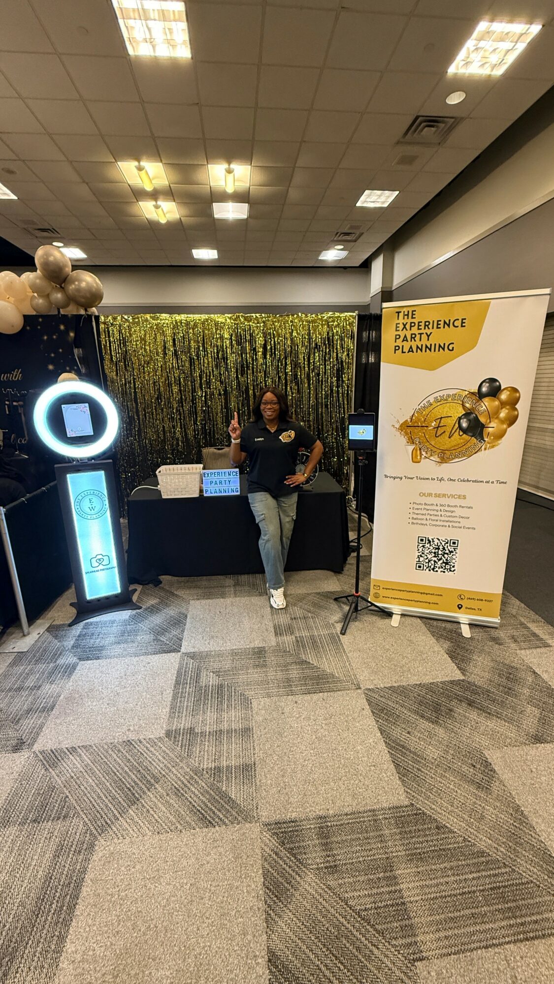 Person standing next to a booth with a yellow and black sign, in an indoor conference setting.