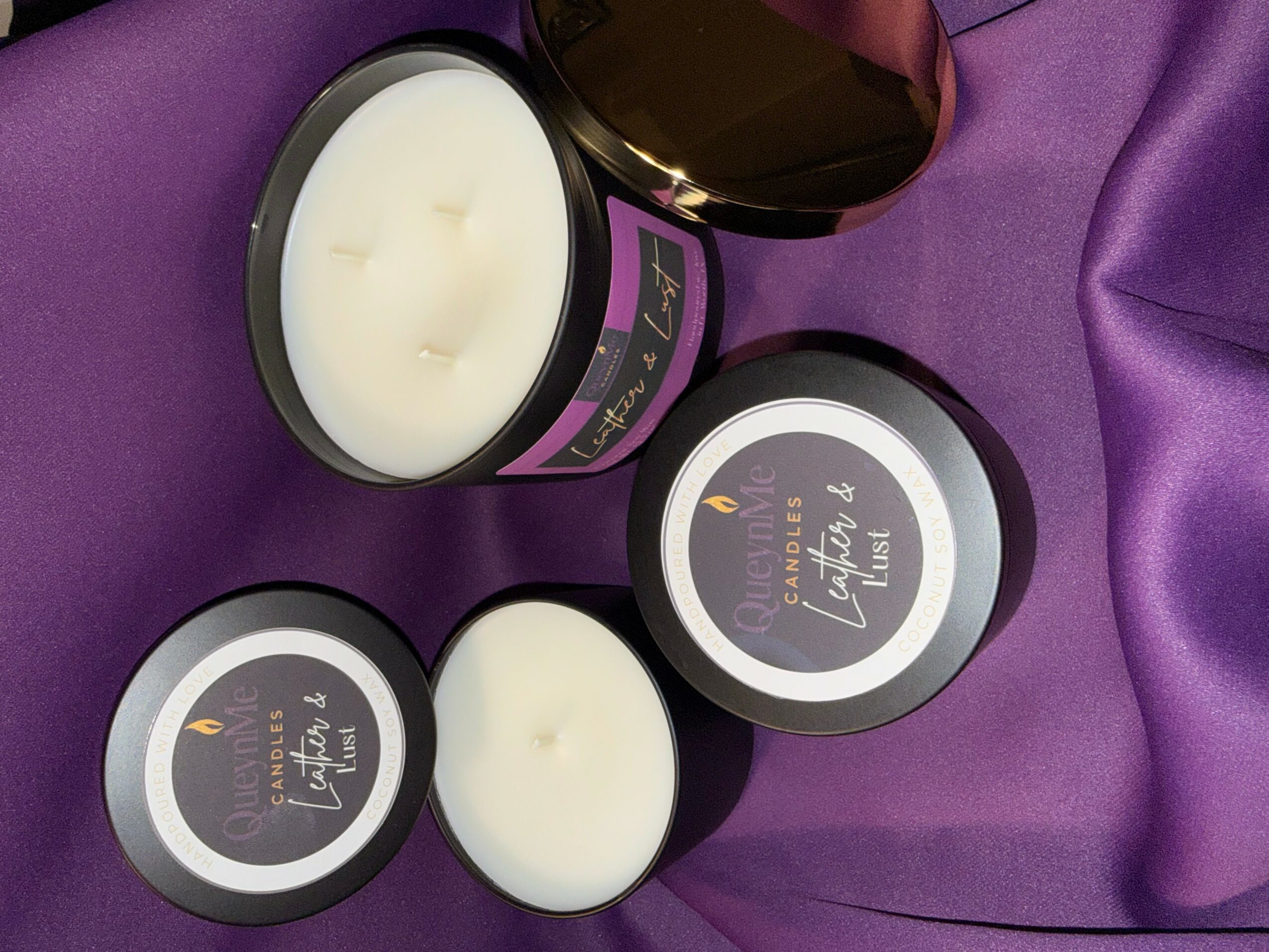 Three black containers with white candles on purple fabric, one open showing the candle inside, and a brown jar in the background.