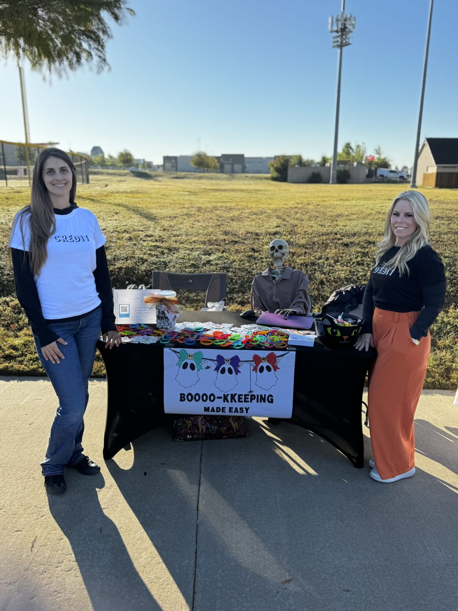 Two women stand beside a table with a skeleton figure, outdoors on a sunny day. The table has a sign and various items.