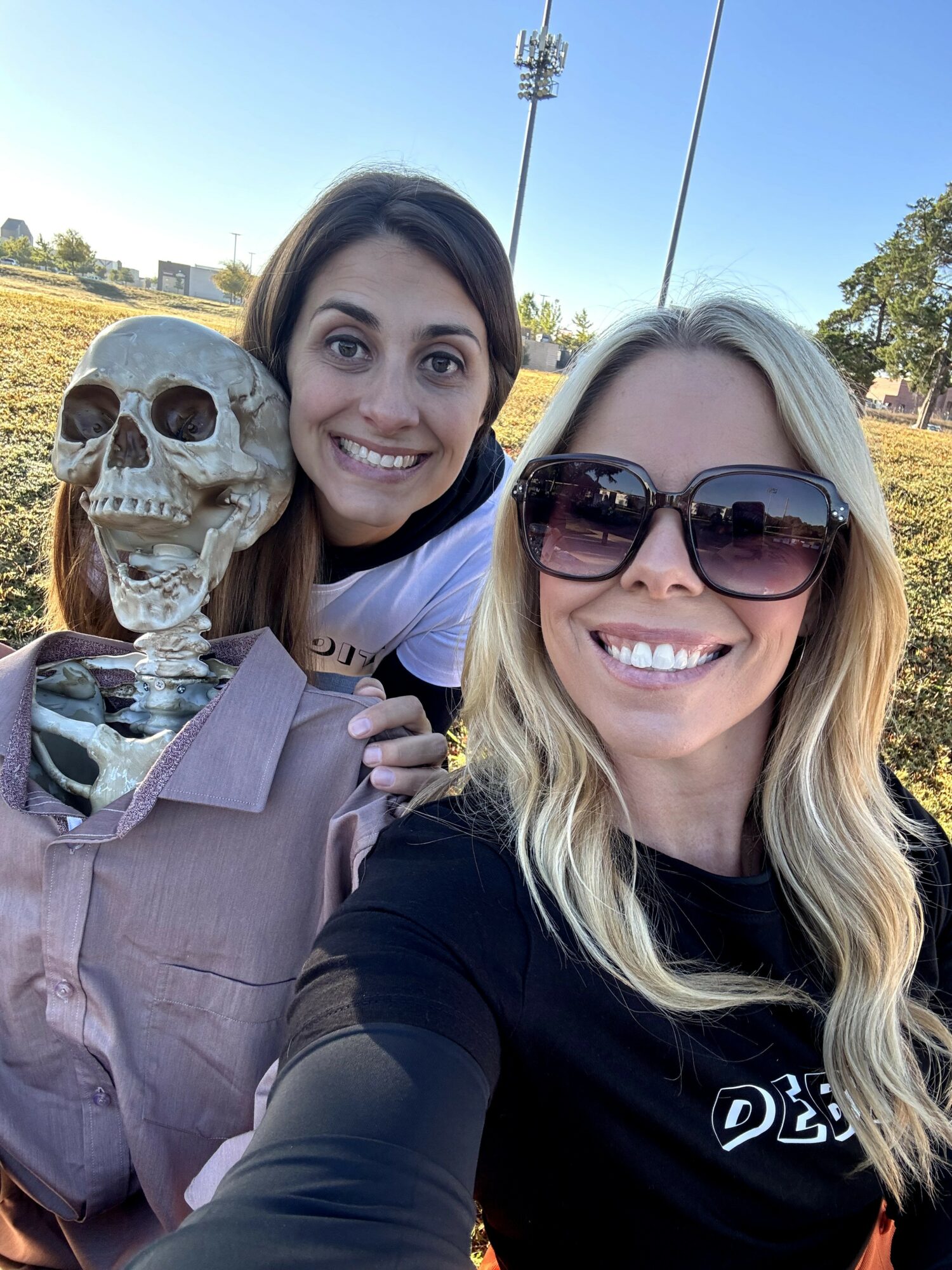Two women smiling outdoors, one holding a skeleton prop, with trees and a clear sky in the background.