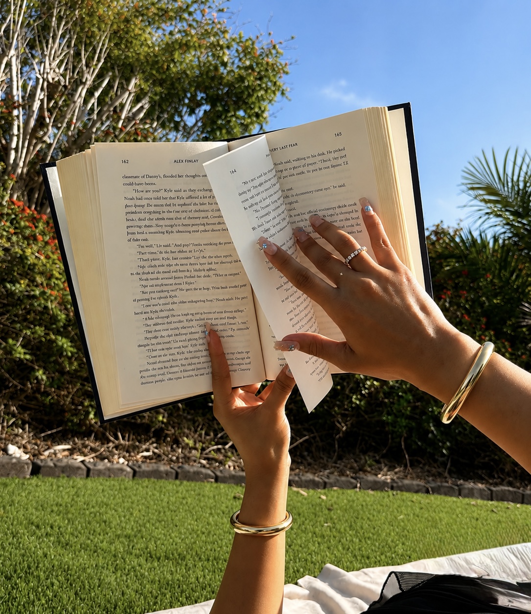 Person holding an open book outdoors with trees and blue sky in background, sunlight illuminating the scene.