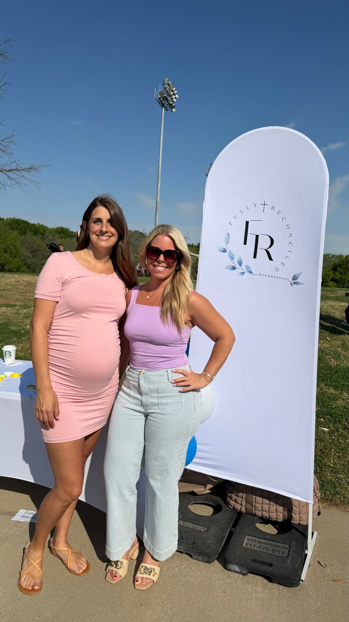 Two women standing outdoors on a sunny day, smiling, with a white backdrop and a tall light pole behind them.