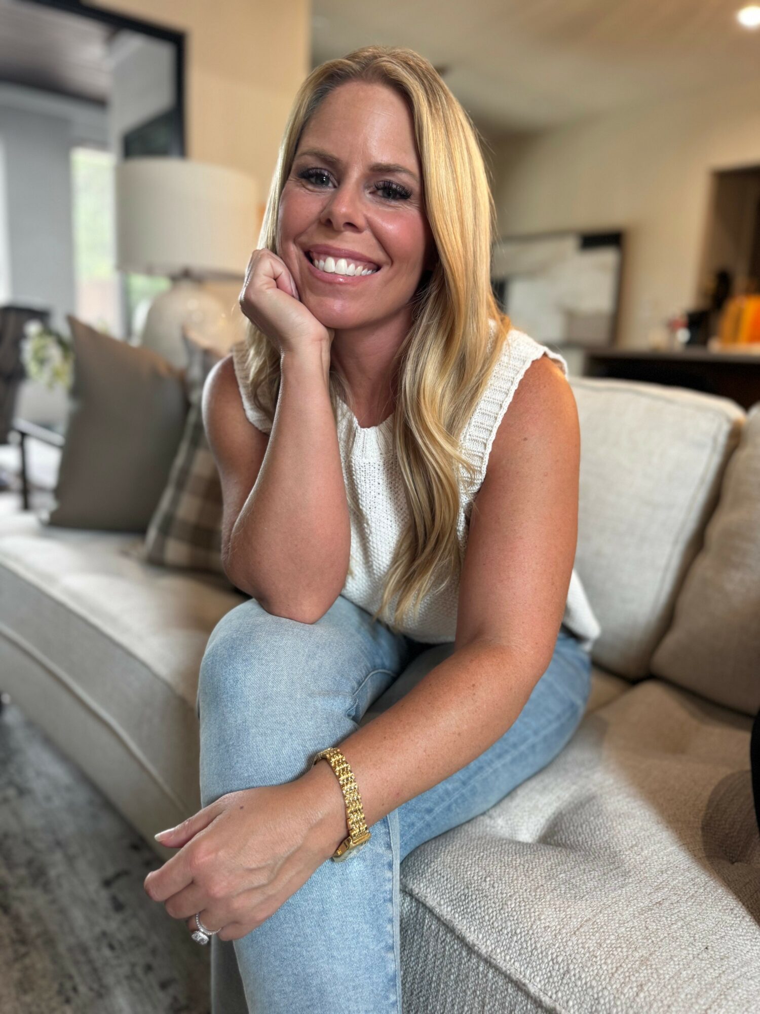 Smiling woman with long blonde hair sitting on a beige sofa in a living room, resting her chin on her hand, wearing a white top and jeans.