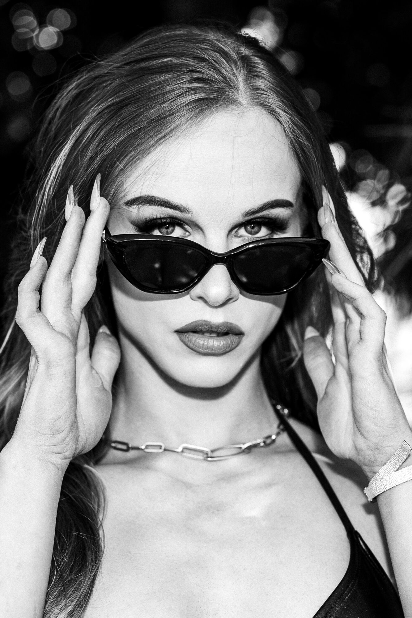 Woman with sunglasses adjusting them, wearing a necklace, with blurred lights in background.