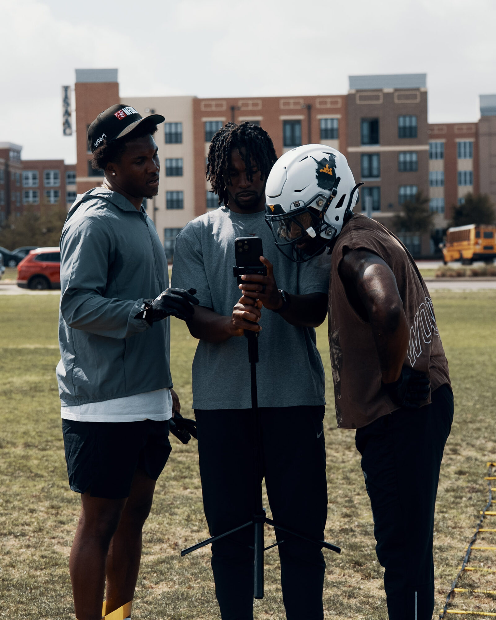 Three people stand outdoors, one wearing a helmet, looking at a phone, with a building and cars in the background.