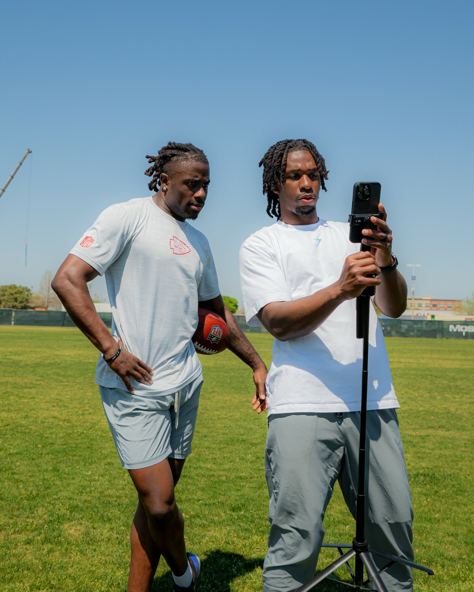 Two men stand on a grassy field, one taking a selfie with a smartphone, both wearing white shirts and shorts, under a clear blue sky.