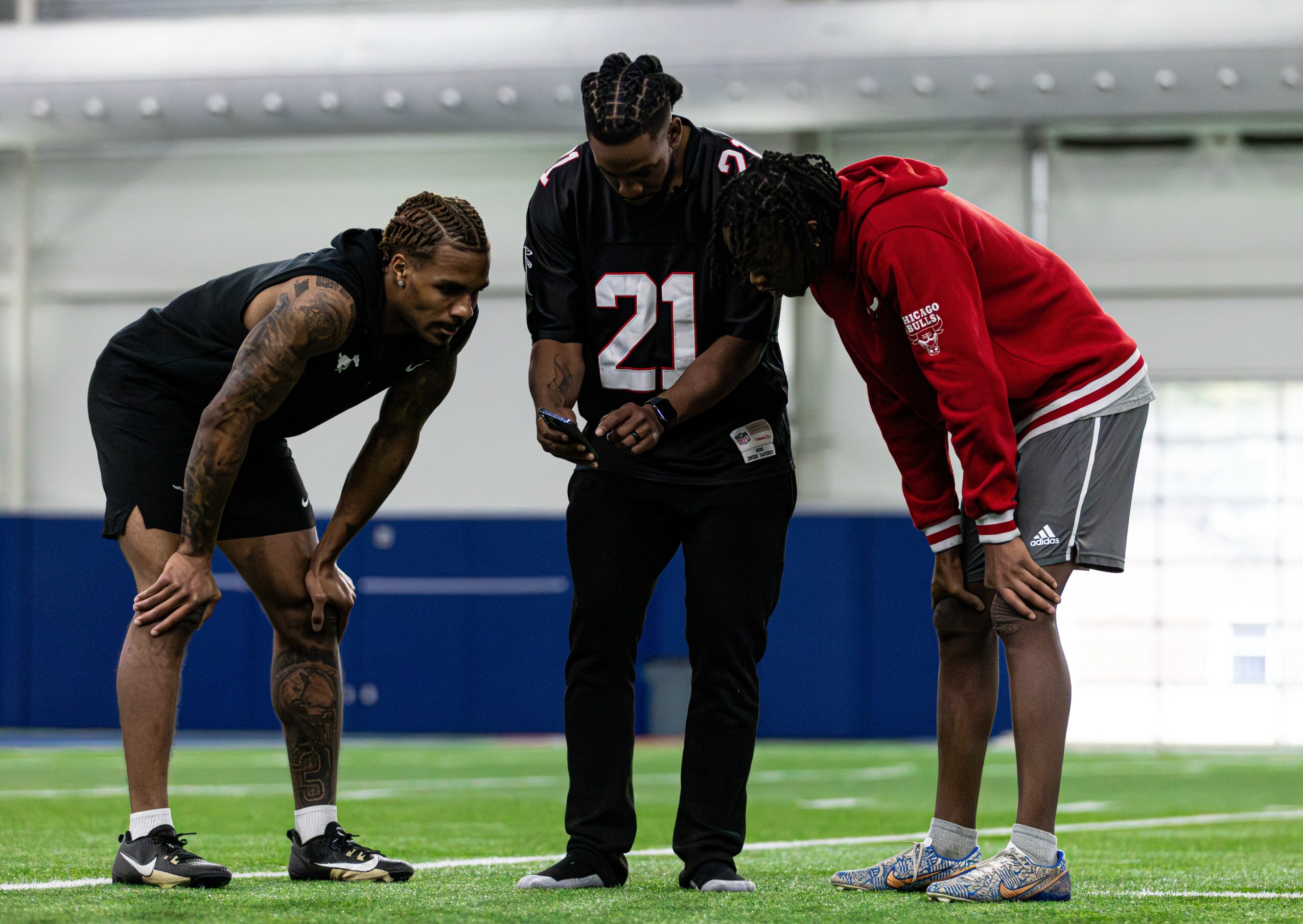 Three people on a sports field, one in a red hoodie, two in black, bending over and looking at something.