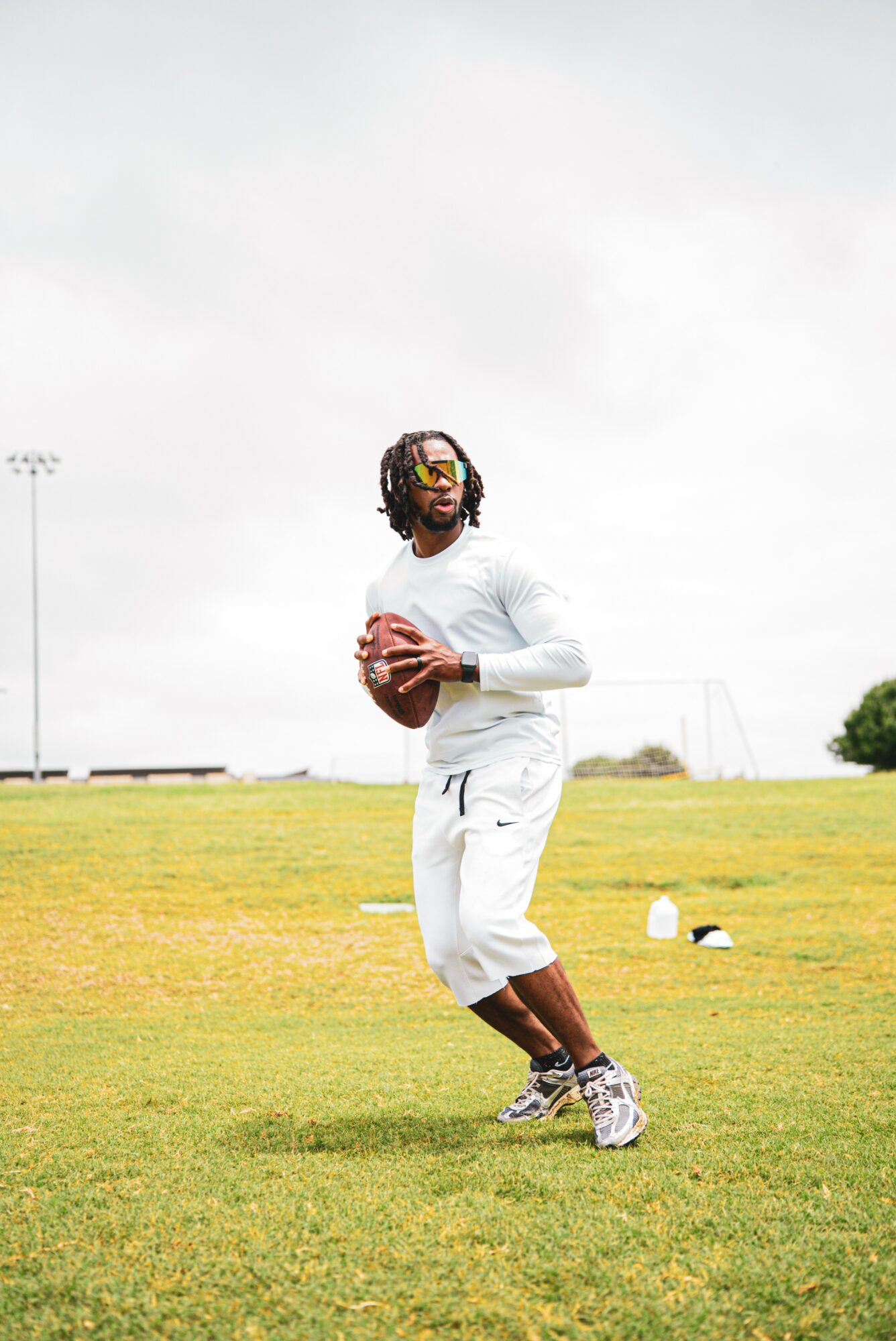 Person in white pants and shirt holding a football on a grassy field, with a cloudy sky and sports field in background.