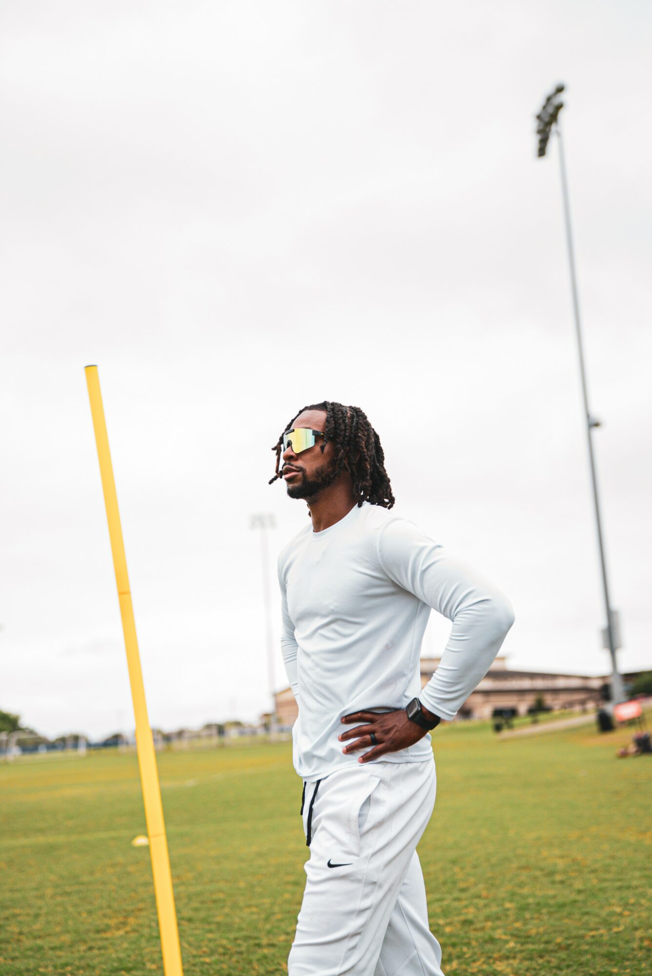 Man with sunglasses and dreadlocks stands on a sports field with hands on hips, wearing a white long-sleeve shirt and pants.