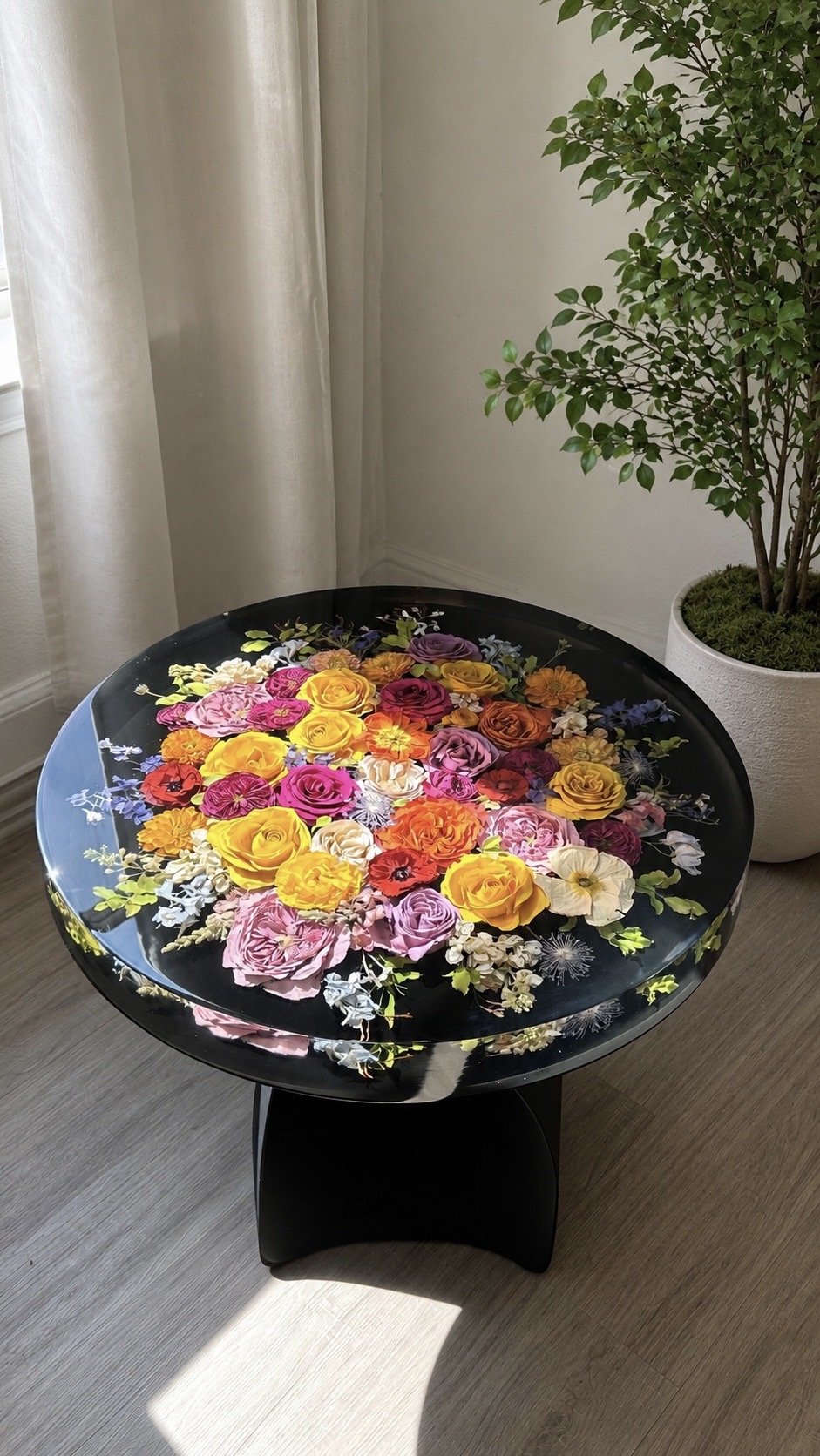 Round black table with a floral arrangement of yellow, pink, orange, and purple flowers, next to a potted plant and curtains.