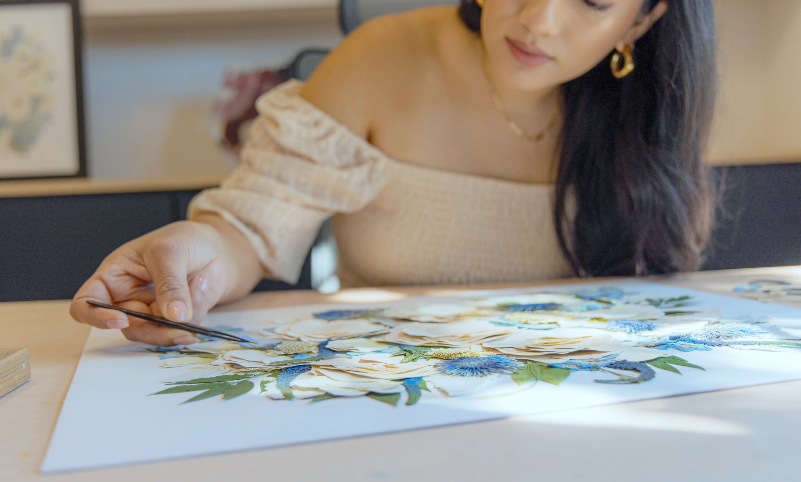 Woman with long dark hair and earrings works on a floral painting at a table.