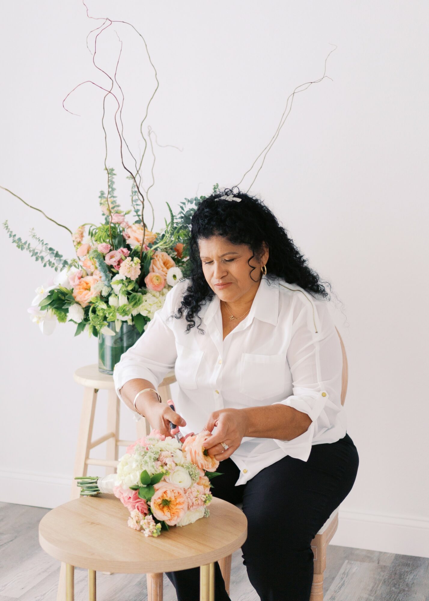 Woman arranging flowers on a small table with a large floral arrangement behind her, in a bright room.