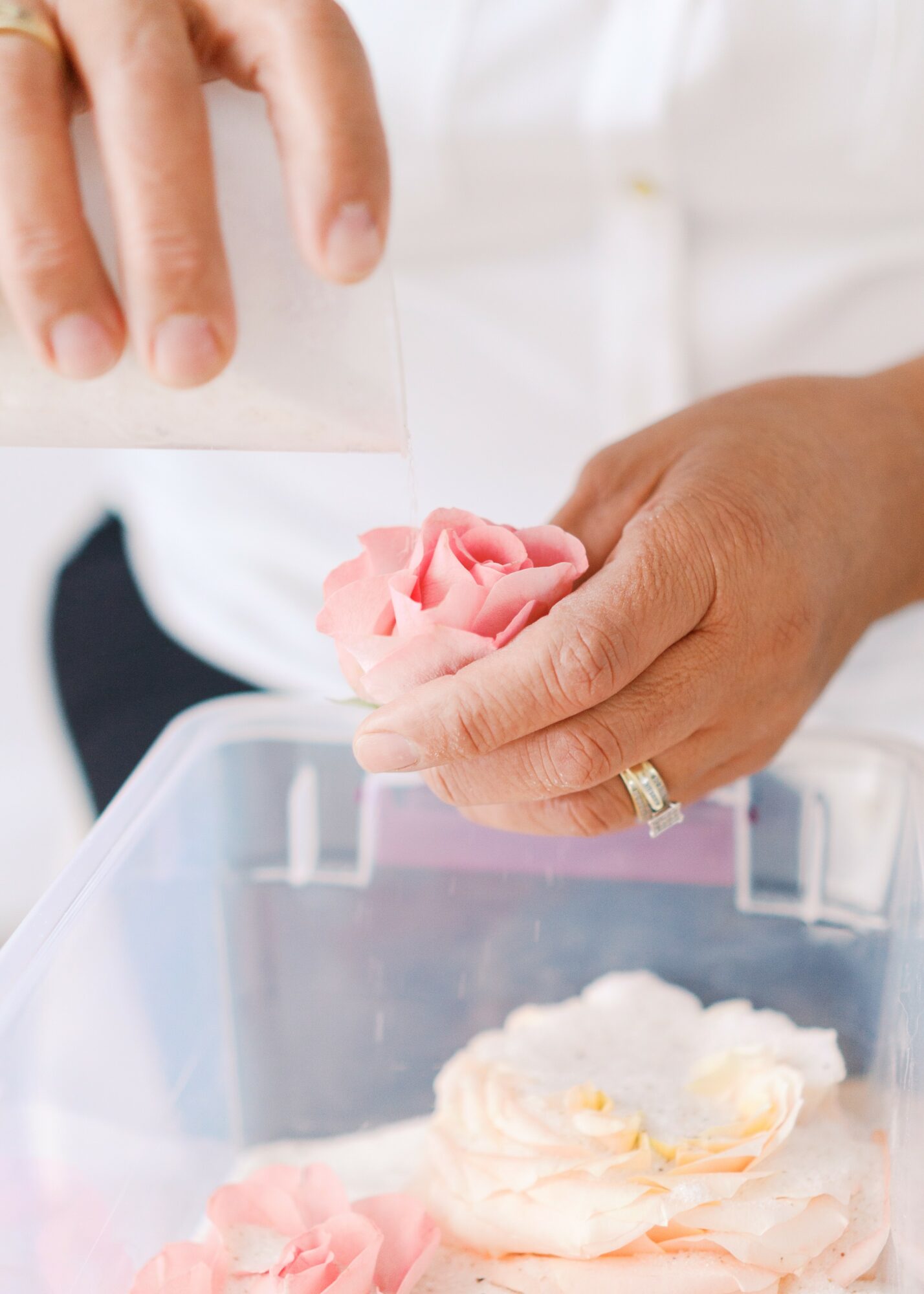Person holding a pink flower-shaped object over a clear container, with a hand wearing a ring, in a bright setting.