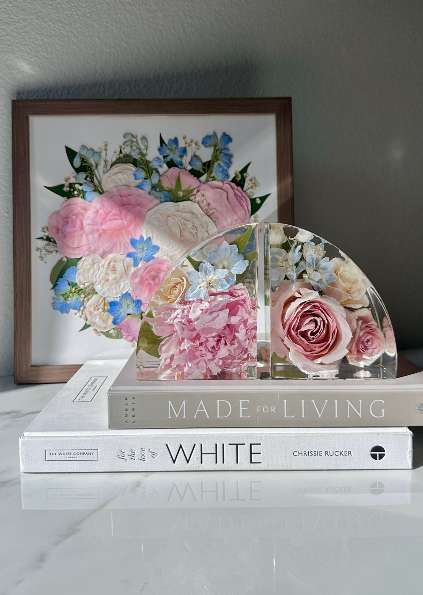 Framed floral artwork with pink, blue, and white flowers, and two glass floral sculptures on books, against a gray wall.