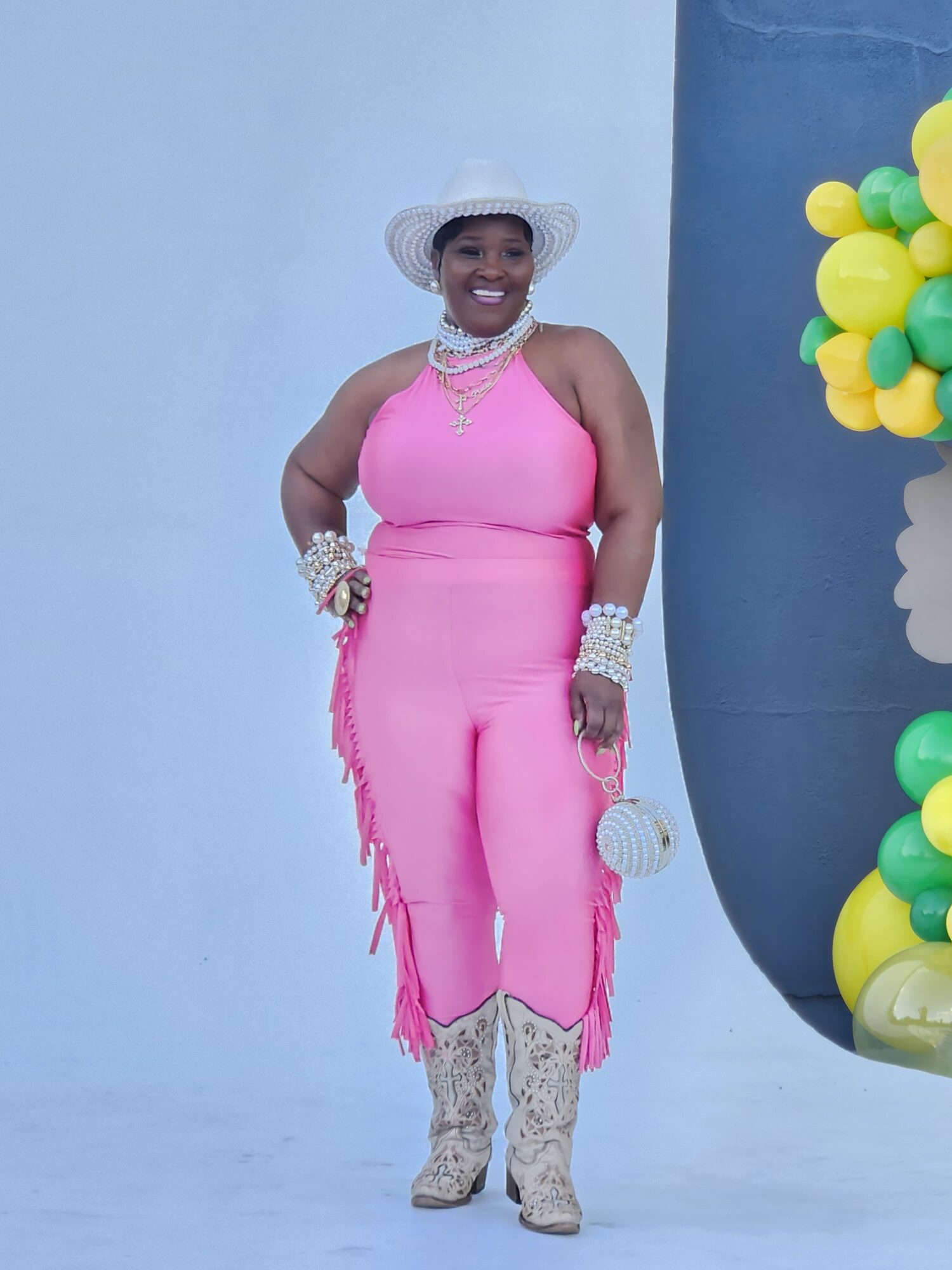 Woman in pink dress with hat, jewelry, and boots, smiling, standing against a light blue background.