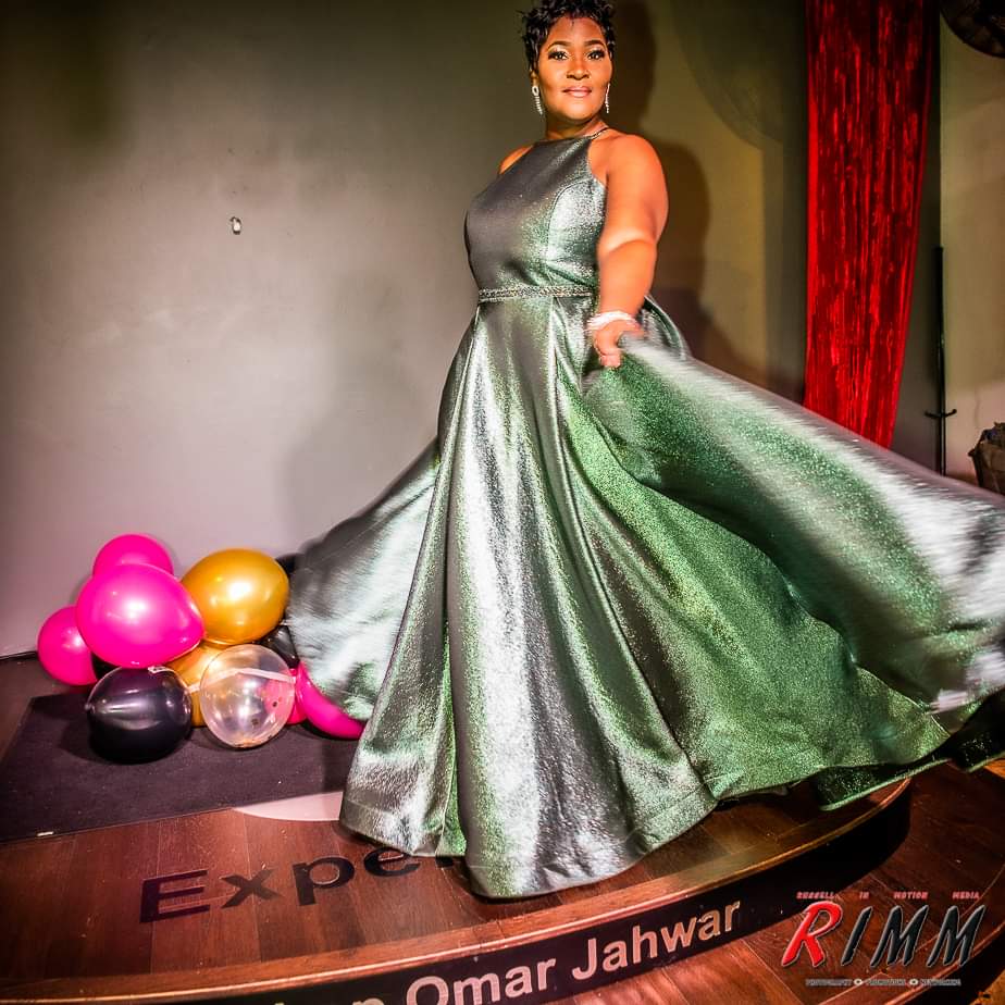 Woman in a shiny green gown standing on stage with balloons nearby, red curtain in background, name and event visible.