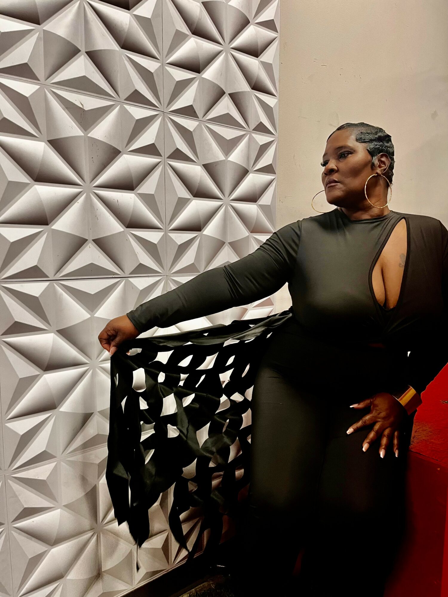 Woman in black dress with cut-out shoulder, standing against a textured wall, holding a black patterned shawl, with her hand on a red surface.