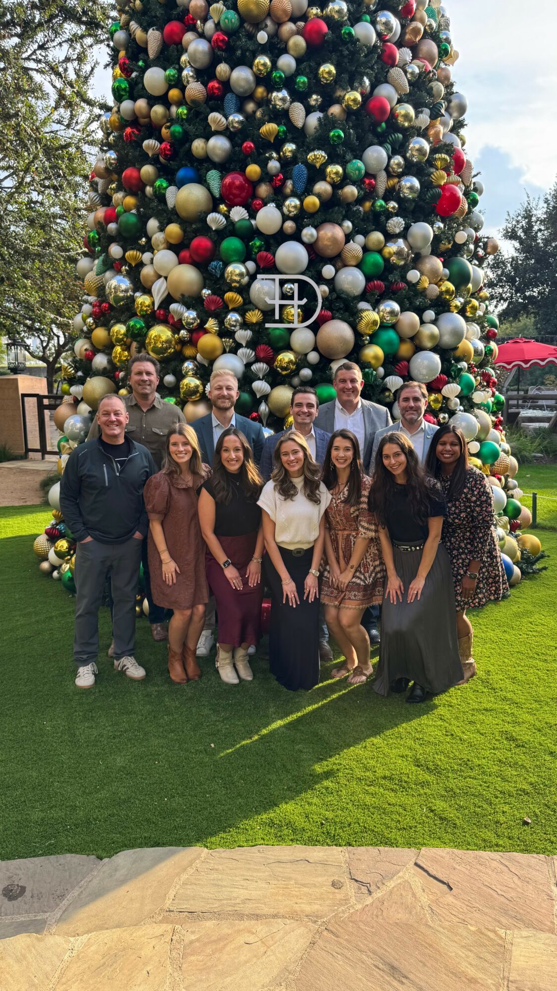 Group of ten people standing in front of a large decorated Christmas tree outdoors, smiling, with grass and trees in background.
