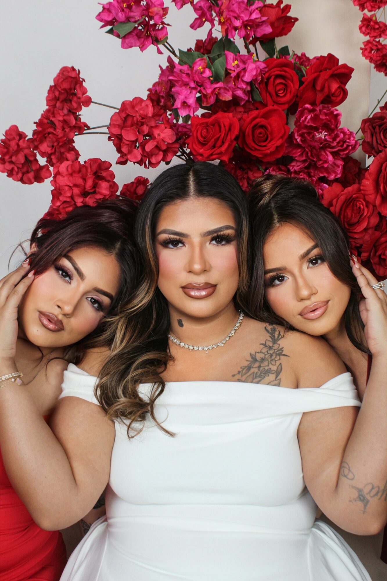 Three women with dark hair and makeup, surrounded by red and pink flowers, posing closely together.