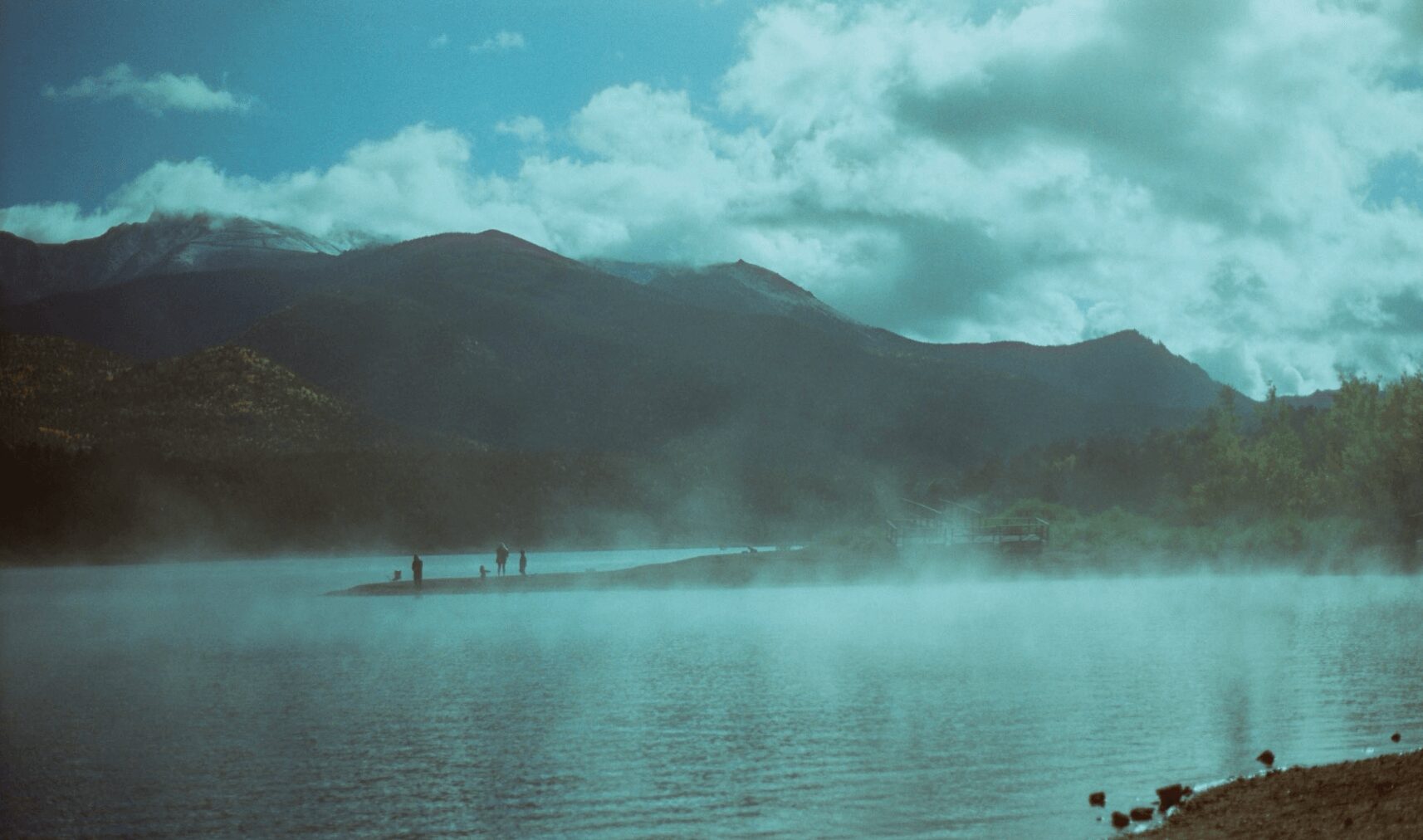 A lake with mist, mountains in the background, and a cloudy sky, with a few people walking along the shore.