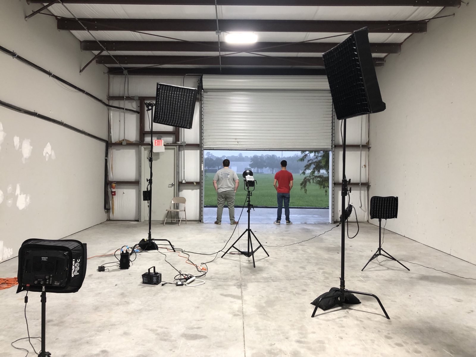 Empty indoor space with lighting, speakers, and tripods, facing an open garage door with three people outside, grass, and trees.