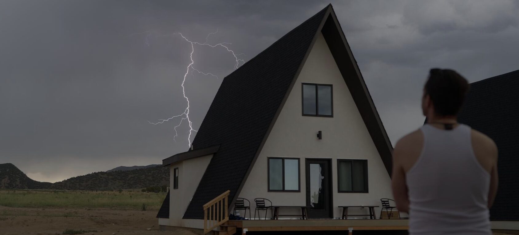 Person with back facing a house during a lightning storm, dark cloudy sky, landscape background.