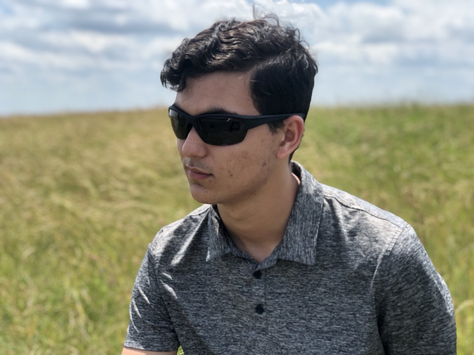 Young man with dark hair wearing sunglasses and a gray shirt outdoors on a grassy field under a partly cloudy sky.