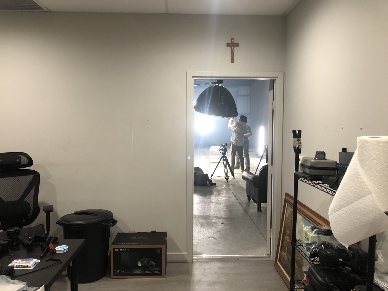 Person taking a photo in a mirror, with a cross on the wall above the doorway, in a room with various objects.