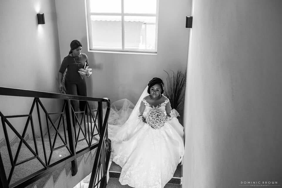 A bride in a wedding dress sits on stairs near a window, with a woman in the background holding a tray.