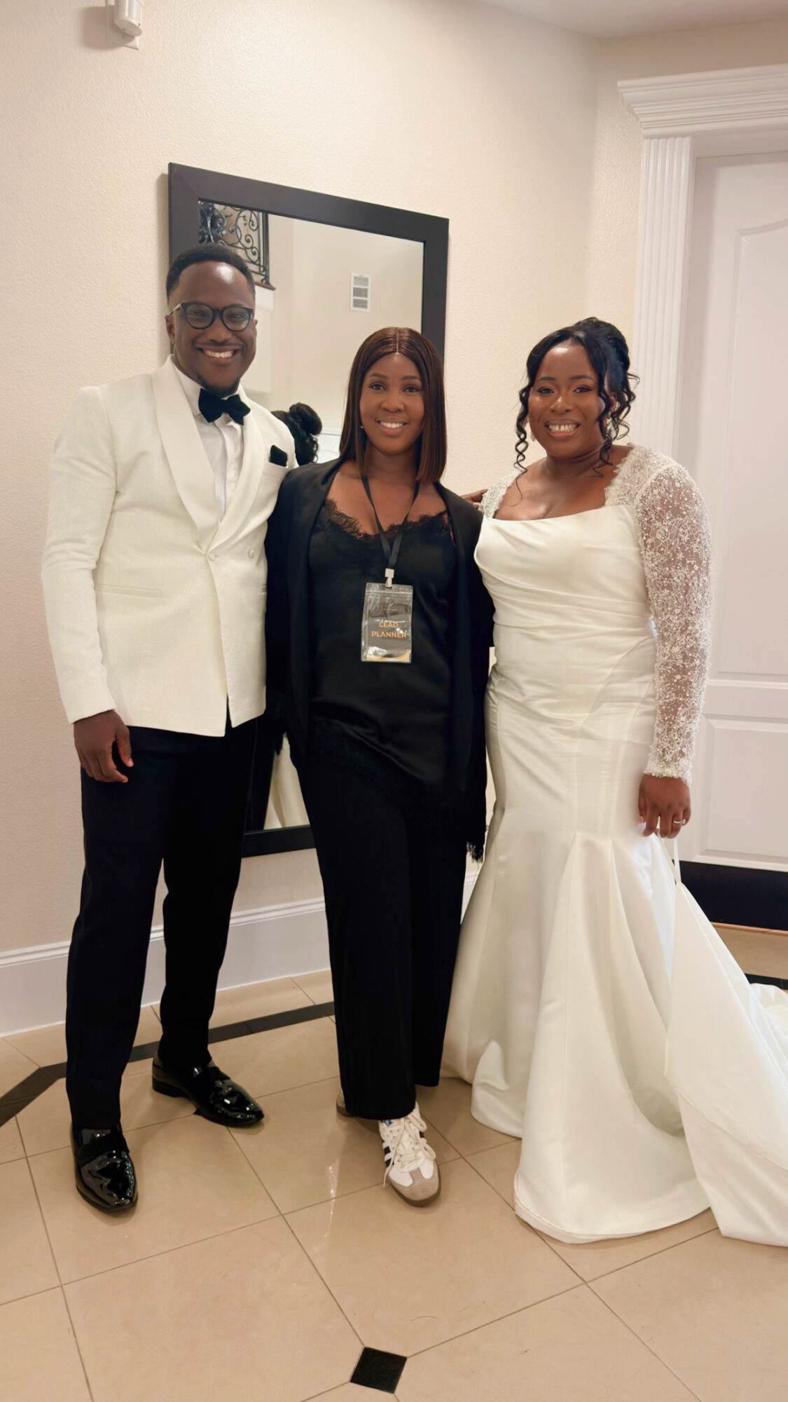 Three people dressed formally, two women and one man, standing indoors in front of a mirror and smiling.