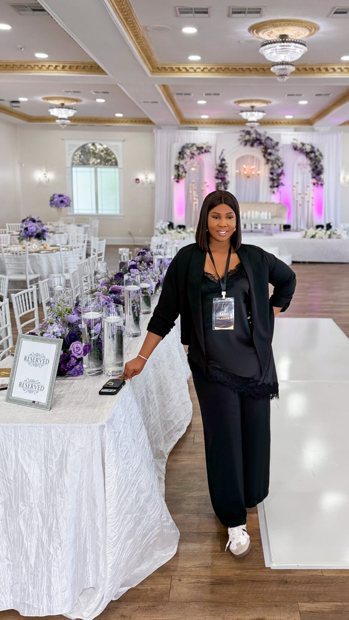 Woman standing in decorated event hall with purple and white floral arrangements, white tables, and elegant ceiling details.