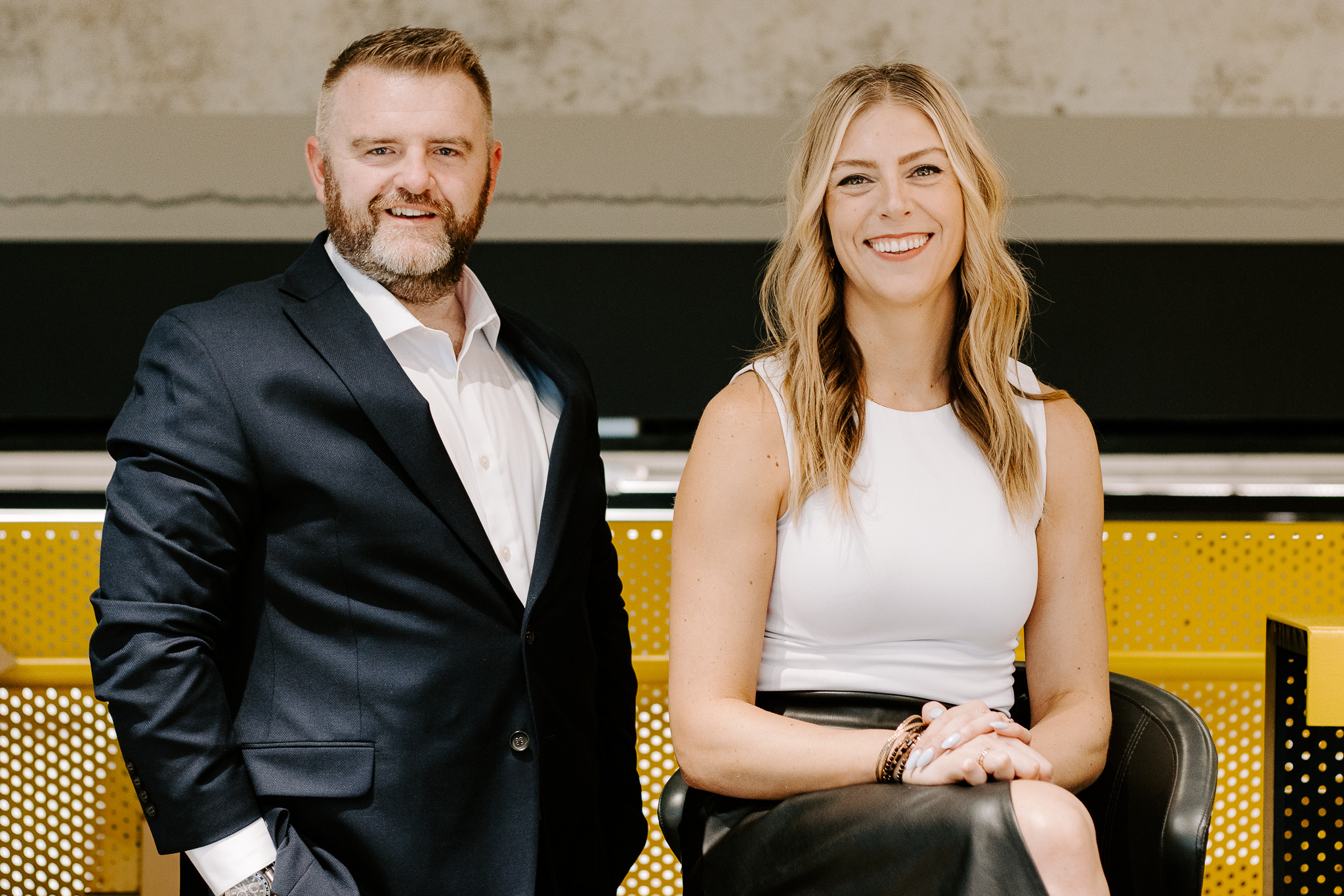 A man and woman stand side by side, smiling, in a professional setting with a yellow and black background.