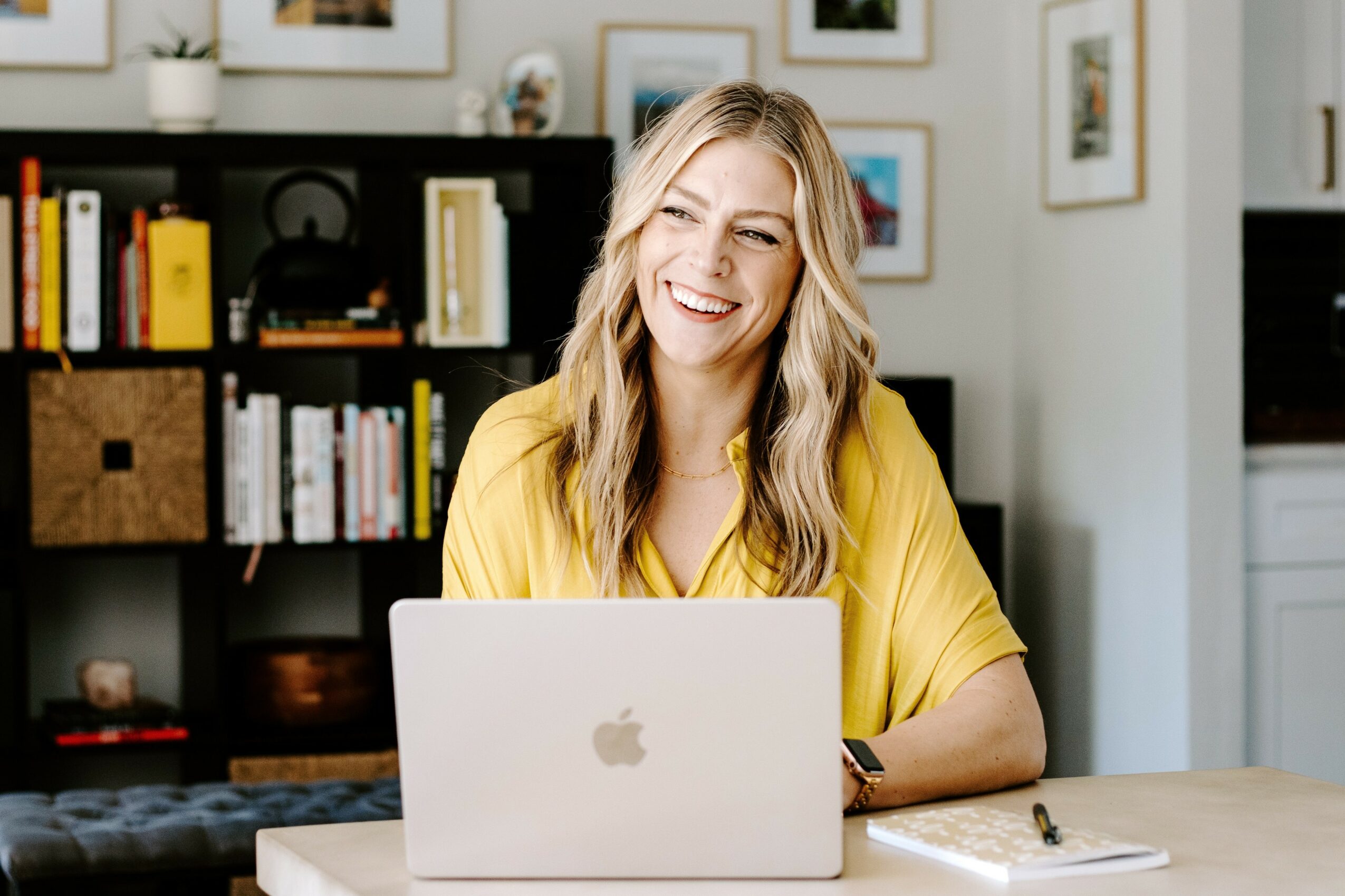 Woman with long blonde hair smiling at a laptop in a bright room with bookshelves.