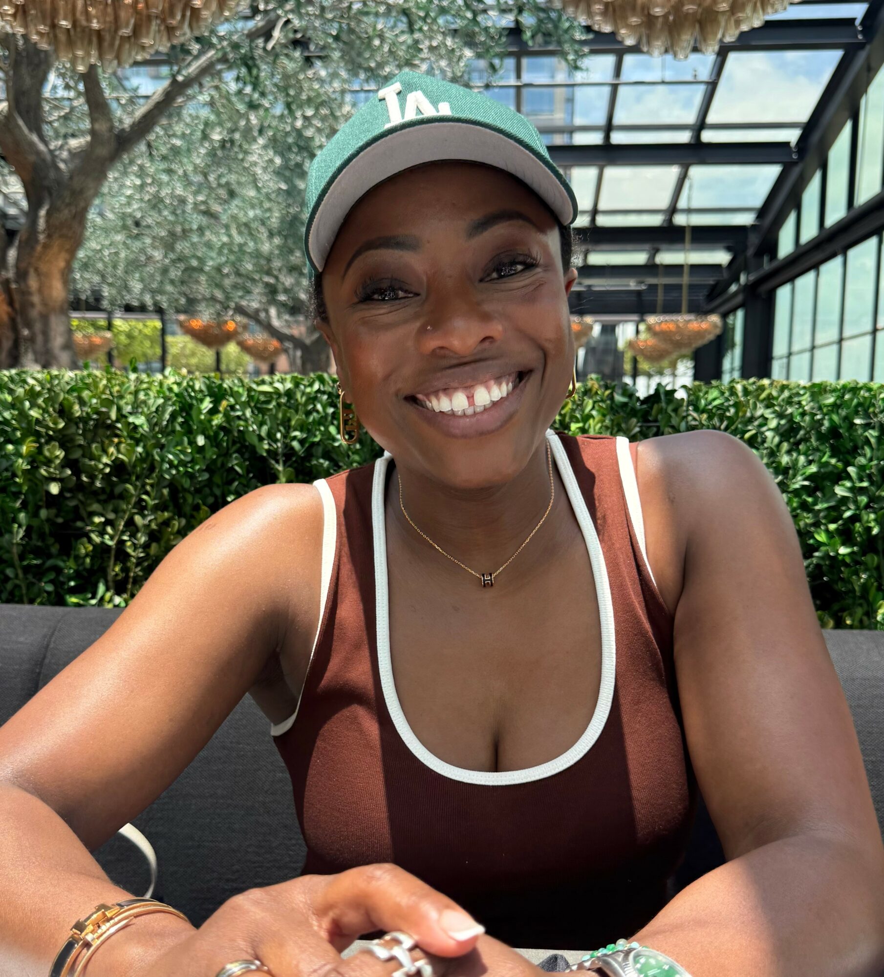 Smiling woman wearing a cap and tank top, sitting outdoors with greenery and glass structure behind her.