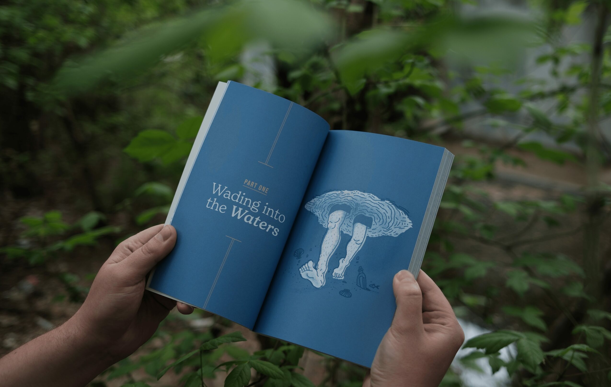 Person holding open book with blue pages, showing an umbrella and water, in a green outdoor setting.