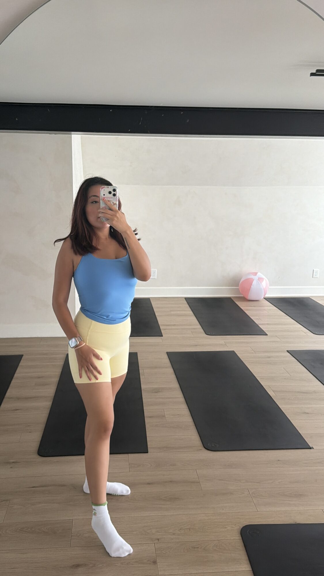 Person taking a mirror selfie in a gym, wearing a blue top and yellow shorts, with black yoga mats on the floor.