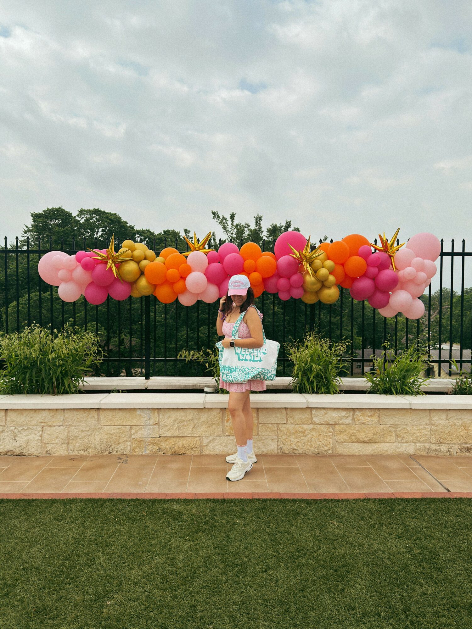 Person holding colorful balloons outdoors near a fence and greenery, cloudy sky in background.