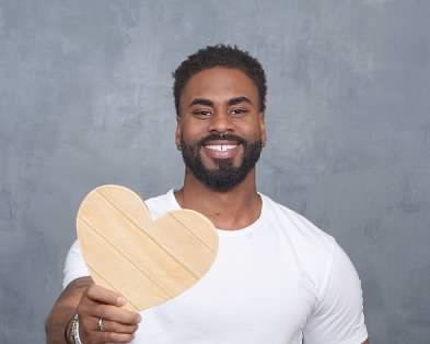 Man with a beard smiling holding a wooden heart-shaped object, wearing a white shirt, against a gray background.